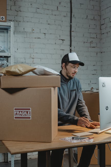 Man working on computer in warehouse with packages. E-commerce shipping.