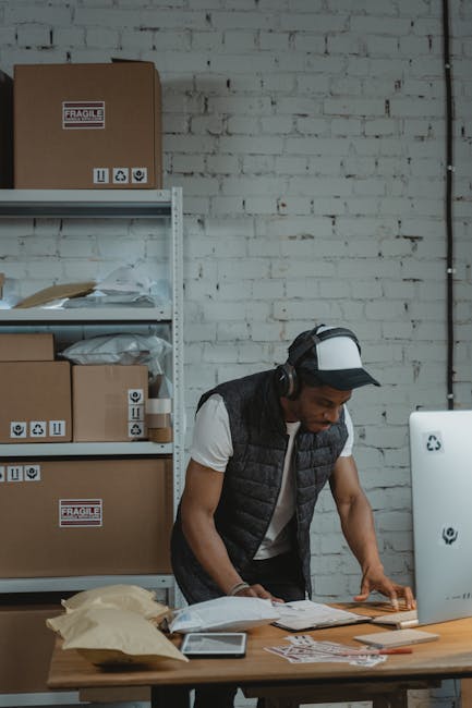 A warehouse worker uses a computer to manage shipments surrounded by parcels and shelves in an industrial setting.